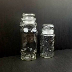May include: Two clear glass jars with lids. The larger jar has a ribbed design and the smaller jar has a diamond pattern.