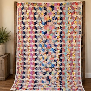 May include: A rectangular quilt featuring a repeating tumbling block pattern composed of numerous small fabric patches in various colors including blue, pink, yellow, and red. The quilt has a white fabric border and a visible stitched texture.