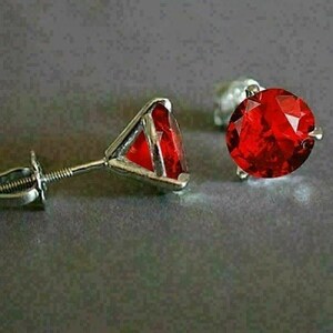 May include: A pair of stud earrings featuring vibrant red gemstones. The gemstones are set in a silver metal setting. One earring is in focus, showing the post and screw back. The other earring is slightly out of focus.