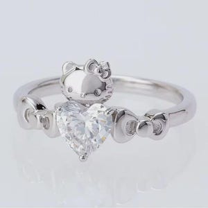 May include: A silver ring with a heart-shaped clear gemstone. The ring features a Hello Kitty design and bow details. The ring is set against a white background. The ring is a piece of jewellery.