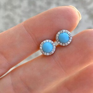May include: A pair of silver stud earrings with a light blue gemstone surrounded by a halo of small clear stones.