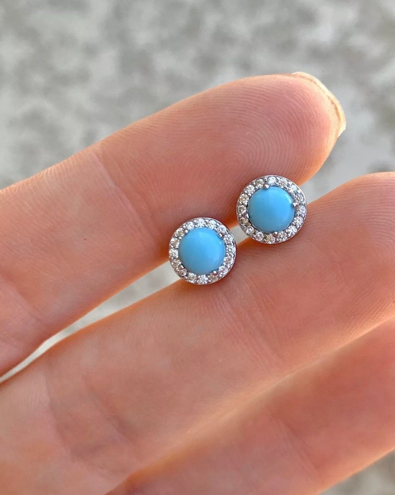 May include: A pair of silver stud earrings with a light blue gemstone surrounded by a halo of small clear stones.