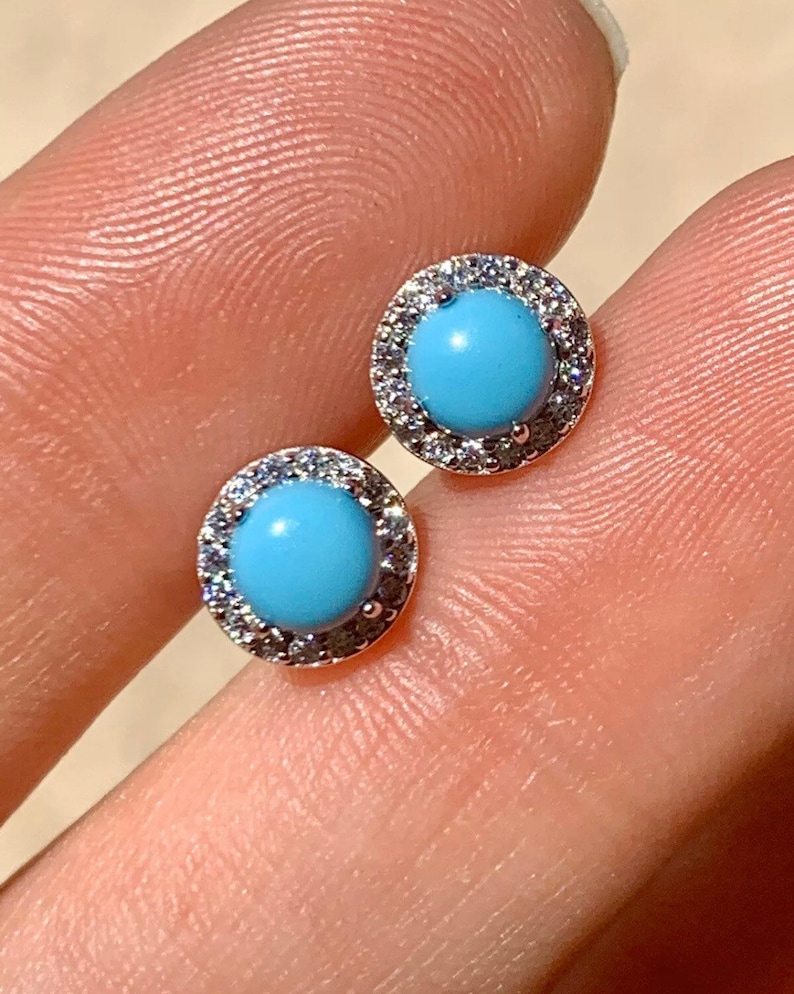May include: A pair of silver earrings with a round blue turquoise stone set in a halo of clear cubic zirconia stones.