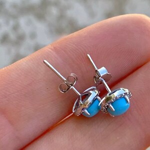 May include: A pair of silver earrings with a turquoise stone set in a halo of smaller stones. The earrings have a post back closure.