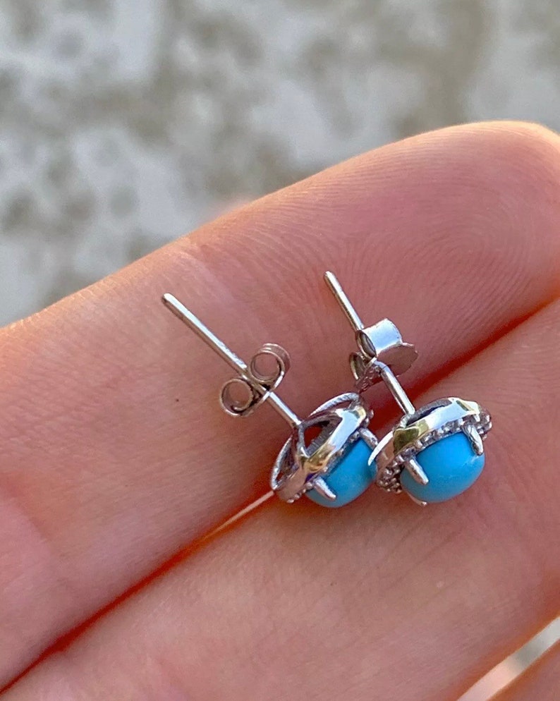 May include: A pair of silver earrings with a turquoise stone set in a halo of smaller stones. The earrings have a post back closure.