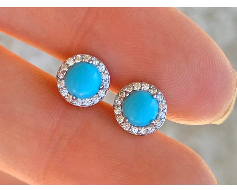 May include: A pair of silver stud earrings with a turquoise stone surrounded by a halo of smaller clear stones.
