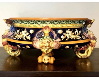 Könnte beinhalten: Eine große, ovale Keramikschale mit blauer und gelber Glasur. Die Schale ist mit aufwendigen floralen und tierischen Motiven in Grün-, Gelb- und Rosatönen verziert. Sie hat drei Beine in Form von stilisierten Tierpfoten.