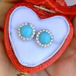 May include: A pair of silver stud earrings with a turquoise stone surrounded by a halo of clear stones. The earrings are in a red heart-shaped box.