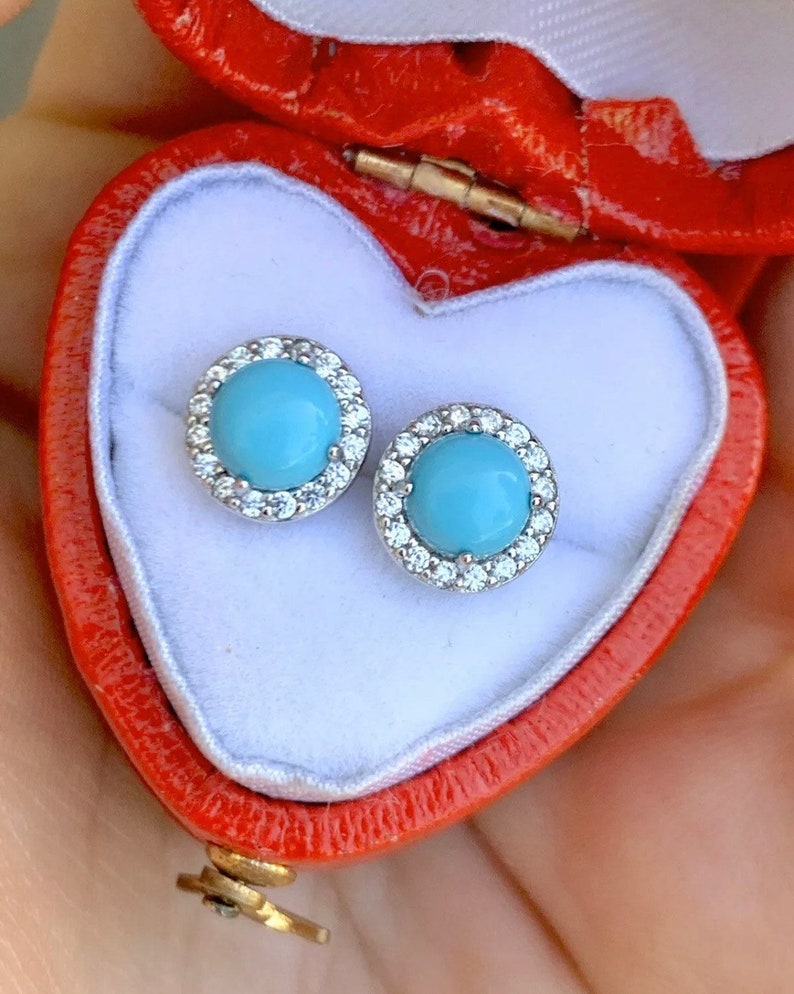 May include: A pair of silver stud earrings with a turquoise stone surrounded by a halo of clear stones. The earrings are in a red heart-shaped box.