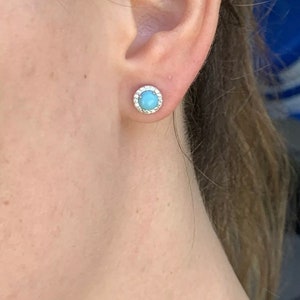 May include: A pair of silver stud earrings with a light blue gemstone surrounded by a halo of smaller white gemstones.