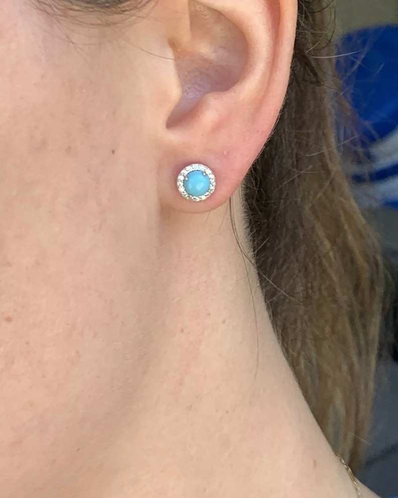 May include: A pair of silver stud earrings with a light blue gemstone surrounded by a halo of smaller white gemstones.