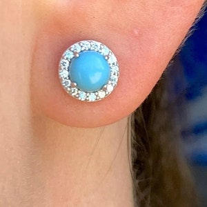 May include: A pair of silver stud earrings with a light blue gemstone surrounded by a halo of clear cubic zirconia stones.
