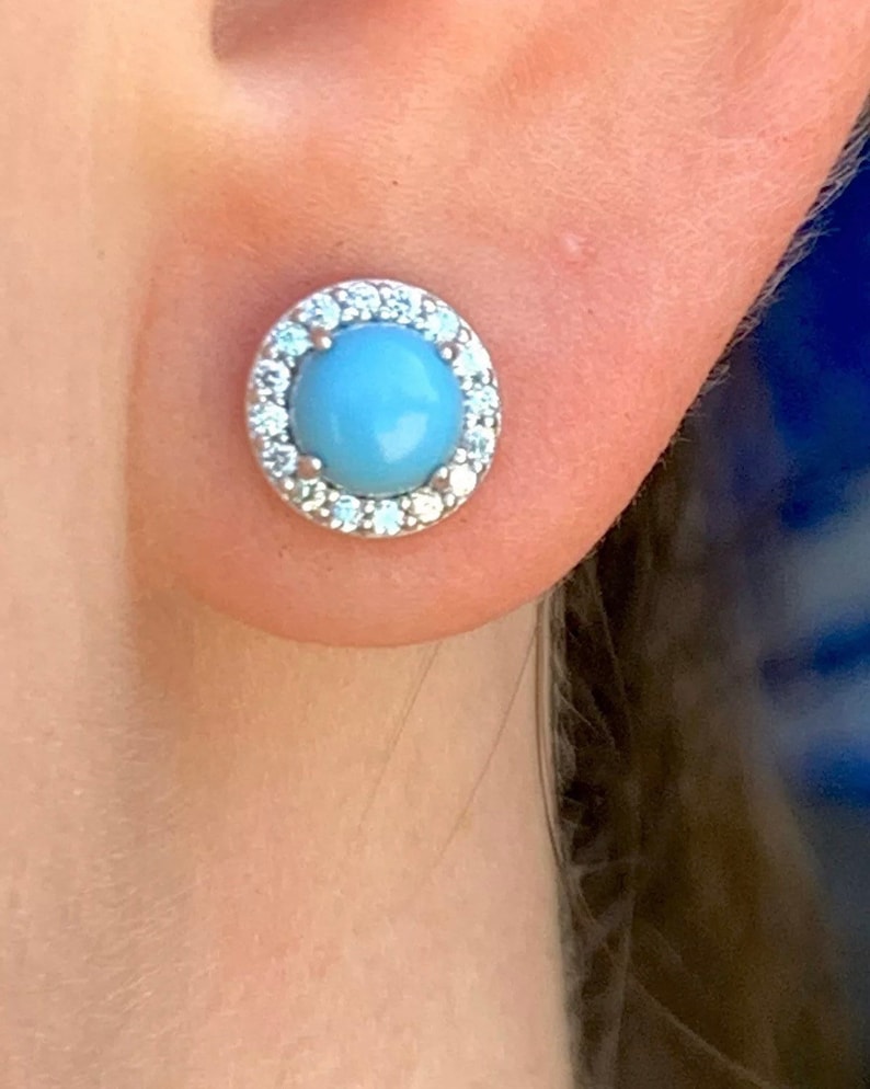 May include: A pair of silver stud earrings with a light blue gemstone surrounded by a halo of clear cubic zirconia stones.