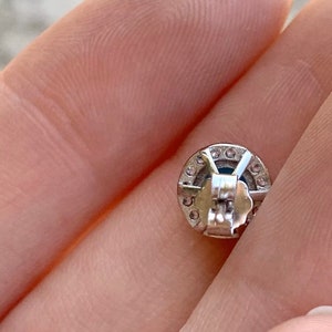 May include: Silver earring with a round, clear stone surrounded by smaller clear stones. The earring has a post back.