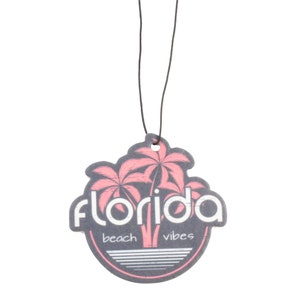 May include: A round car air freshener with a black background and pink palm trees. The text "Florida beach vibes" is printed on the air freshener.