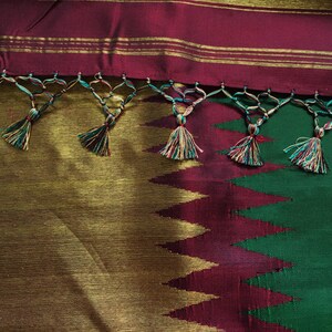 May include: A green and burgundy silk saree with gold trim and tassels. The saree has a geometric pattern and is decorated with gold thread.