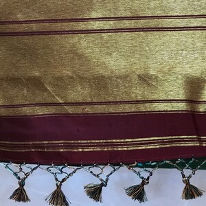 May include: Close-up of the bottom edge of a green and gold sari with a maroon border and gold trim. The sari has a fringe with tassels.