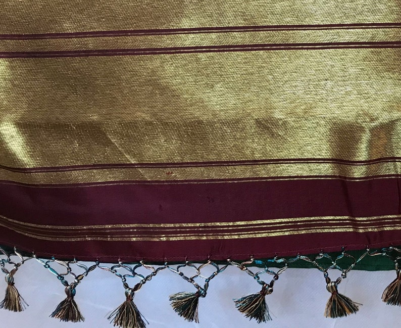 May include: Close-up of the bottom edge of a green and gold sari with a maroon border and gold trim. The sari has a fringe with tassels.