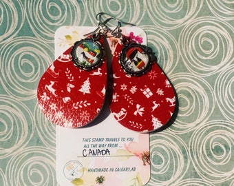 Puede incluir: Pendientes rojos en forma de lágrima con un patrón navideño festivo y ganchos plateados. Cada pendiente presenta un pequeño dije redondo con una colorida escena invernal. Los pendientes están en una etiqueta que dice "CANADA" y "Handmade in Calgary, AB."