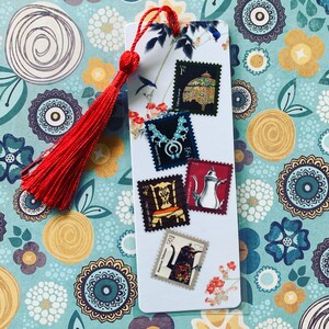 Puede incluir: Un marcapáginas blanco con una borla roja, adornado con sellos postales vintage. Los sellos presentan varios diseños, incluyendo una lámpara Tiffany, un collar turquesa y una cafetera plateada. Ilustraciones florales y de pájaros decoran los bordes del marcapáginas.