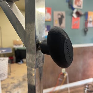 May include: A black knob with the text "Better Studio" attached to a silver metal frame. The frame is square. The background is blurred, showing a room with artwork and a wooden floor.