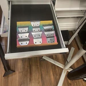 Könnte beinhalten: Eine Schublade in einem weißen Schrank ist geöffnet und zeigt eine Sammlung von Metall-Clippern. Die Klingen sind in einem Regenbogenfarbenen Halter angeordnet. Die Klingen sind in verschiedenen Größen und Stilen.