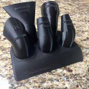 May include: A black 3D-printed stand holding five black funnels of varying shapes and sizes. The funnels have text that reads "BreierStudio" and other text. The stand also has the text "BreierStudio".