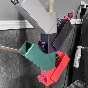 Pode incluir: Um suporte de ferramentas impresso em 3D multicolorido com quatro compartimentos. O suporte é montado em uma haste de metal. Os compartimentos são cinza, azul esverdeado, roxo e vermelho. O suporte é projetado para conter ferramentas como uma broca pequena, um conjunto de pinças e uma serra pequena.