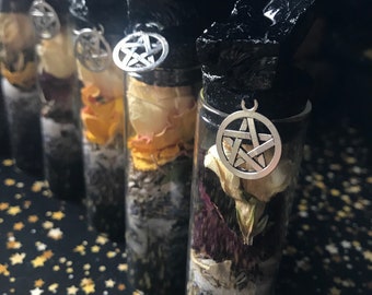 Op de afbeelding: Een rij glazen flessen gevuld met gedroogde bloemen en kruiden. Elke fles heeft een zwarte dop en een zilveren pentagrambedel. De flessen staan op een zwarte ondergrond met gouden sterren.