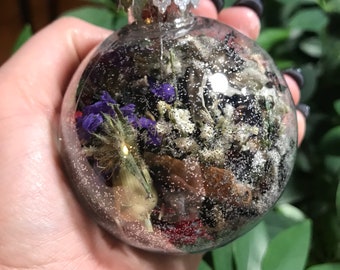 Op de afbeelding: Transparante glazen kerstbal gevuld met gedroogde bloemen, bladeren en glitter. De ornament heeft een zilveren metalen dop en een lus om op te hangen. De bloemen zijn paars, wit en rood.