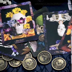Op de afbeelding: Een verzameling tarotkaarten en goudkleurige munten met schedelafbeeldingen. De kaarten tonen bloemstukken met schedels en skeletfiguren. De tekst "The Fool" en "The Hanged Man" is zichtbaar. De munten hebben een schedelontwerp.