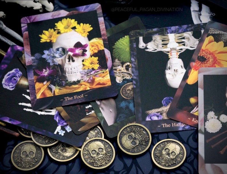Op de afbeelding: Een verzameling tarotkaarten en goudkleurige munten met schedelafbeeldingen. De kaarten tonen bloemstukken met schedels en skeletfiguren. De tekst "The Fool" en "The Hanged Man" is zichtbaar. De munten hebben een schedelontwerp.