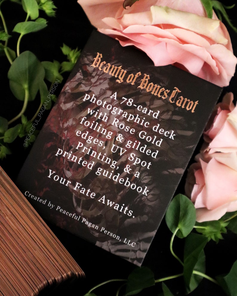 Op de afbeelding: Een zwarte tarotkaartdoos met de titel "Beauty of Bones Tarot" in gouden letters. De doos is omgeven door roze rozen en groen gebladerte. De tekst op de doos beschrijft een fotografisch deck van 78 kaarten met ros&eacute;goudfolie en vergulde randen.