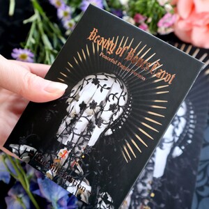 Op de afbeelding: Een zwarte tarotkaartdoos met de titel "Beauty of Bones Tarot" in gouden letters. De doos heeft een illustratie van een schedel versierd met kant en een sunburst-ontwerp. De tekst "Peaceful Pagan Version" is ook zichtbaar.