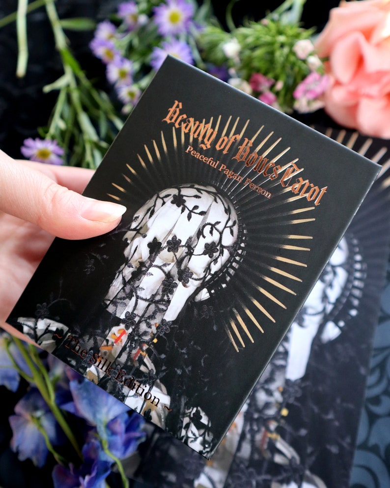 Op de afbeelding: Een zwarte tarotkaartdoos met de titel "Beauty of Bones Tarot" in gouden letters. De doos heeft een illustratie van een schedel versierd met kant en een sunburst-ontwerp. De tekst "Peaceful Pagan Version" is ook zichtbaar.