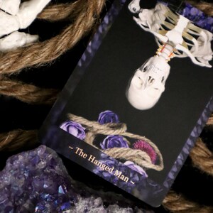 Op de afbeelding: Een tarotkaart met een ondersteboven hangend skelet, versierd met een touw en paarse rozen. De kaart draagt de titel "The Hanged Man" en staat tegen een zwarte achtergrond. Amethistkristallen en touw zijn ook zichtbaar.