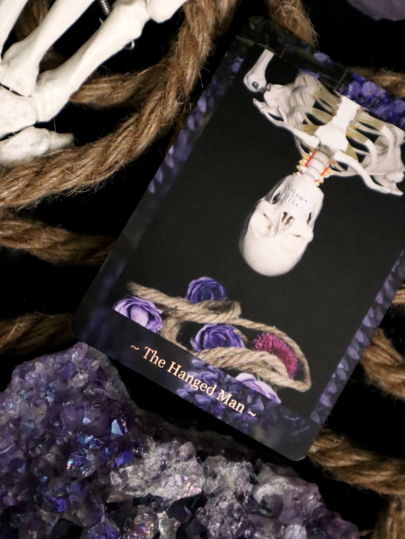 Op de afbeelding: Een tarotkaart met een ondersteboven hangend skelet, versierd met een touw en paarse rozen. De kaart draagt de titel "The Hanged Man" en staat tegen een zwarte achtergrond. Amethistkristallen en touw zijn ook zichtbaar.