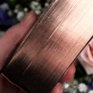 Op de afbeelding: Close-up van een ros&eacute;goudkleurig boek met getextureerde randen. De pagina's van het boek zijn zichtbaar en de rug heeft een metallic glans. De achtergrond toont vage bloemen in tinten roze, wit en blauw.