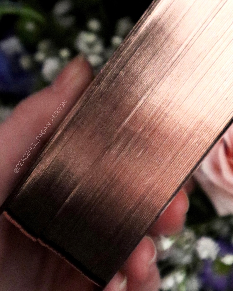 Op de afbeelding: Close-up van een ros&eacute;goudkleurig boek met getextureerde randen. De pagina's van het boek zijn zichtbaar en de rug heeft een metallic glans. De achtergrond toont vage bloemen in tinten roze, wit en blauw.