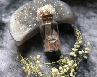 Op de afbeelding: Een kleine glazen fles met een kurk, gevuld met roze en heldere kristallen. De fles is omgeven door een krans van witte bloemen en rust voor een grijze, met kristallen beklede geode.