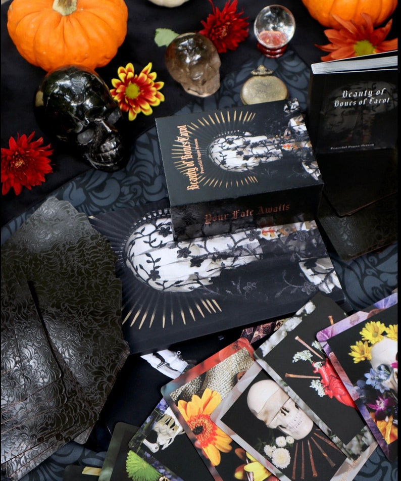 Op de afbeelding: Een gothic stilleven met een zwarte schedel, tarotkaarten en een doos met de tekst "Beauty of Bones of Tarot". De kaarten tonen bloemen- en schedelafbeeldingen. Oranje pompoenen en bloemen zijn verspreid over de sc&egrave;ne, wat een duistere sfeer cre&euml;ert.