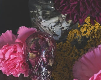 Op de afbeelding: Een heldere hars schedel gevuld met gedroogde rozenblaadjes, omringd door roze anjers, gele bloemen en een donkerpaarse chrysant. Een glazen pot met witte salie staat op de achtergrond. De opstelling staat op een donker houten oppervlak.