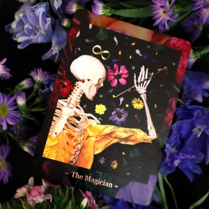 Op de afbeelding: Een tarotkaart met een skelet in een gele jas, tegen een zwarte achtergrond met bloemen en een oneindigheidssymbool. De kaart heet "The Magician" en is omgeven door paarse en roze bloemen.