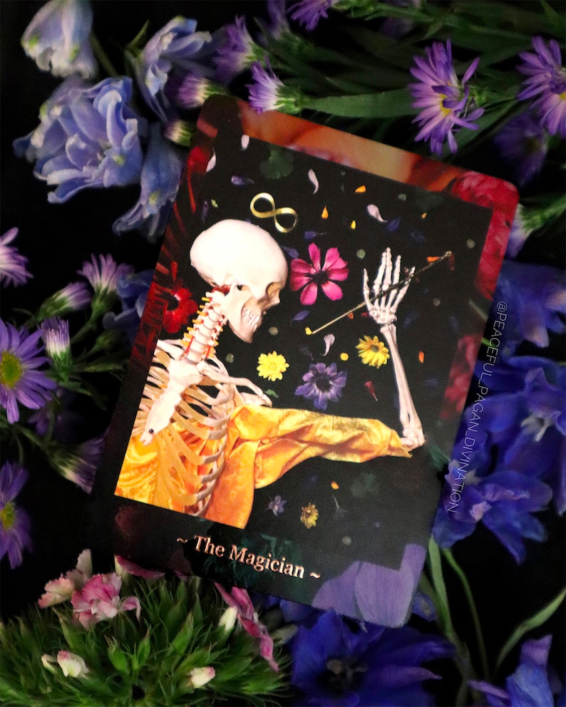 Op de afbeelding: Een tarotkaart met een skelet in een gele jas, tegen een zwarte achtergrond met bloemen en een oneindigheidssymbool. De kaart heet "The Magician" en is omgeven door paarse en roze bloemen.