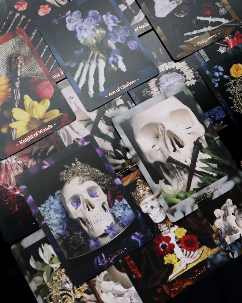 Op de afbeelding: Een verzameling tarotkaarten met donkere achtergronden en gotische beelden. De kaarten bevatten illustraties van schedels, bloemen en skeletachtige handen. Sommige kaarten hebben tekst zoals "Knight of Wands" en "Ace of Chalices".