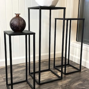 May include: Set of three black metal plant stands. Each stand has a square top and a rectangular frame. A dark brown vase sits on the smallest stand. A white geometric pot with greenery sits on the tallest stand.