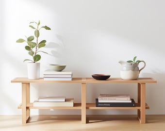 May include: A light-colored wooden bench with two shelves, holding a variety of items. A potted plant, books, and ceramic bowls are arranged on the top shelf. A small pitcher and more books are also visible.