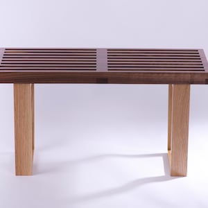 Op de afbeelding: Houten bank met een latten bovenkant in donkerbruin en vier lichtgekleurde houten poten. De bank heeft een minimalistisch ontwerp, met een bovenkant van ongeveer 100 cm lang. Geschikt voor diverse interieurs.