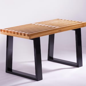 May include: A rectangular bench featuring a slatted light-toned wooden seat with visible grain patterns. The seat is supported by two sturdy, black-painted metal legs that have a wide, angular, U-shaped design, providing a stable base.