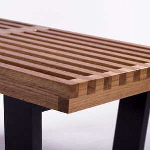 Puede incluir: Banco de madera con tapa de listones y patas negras. El banco es de madera marrón claro con una serie de listones espaciados uniformemente en la parte superior. Las patas son de color negro sólido, lo que contrasta con la madera.